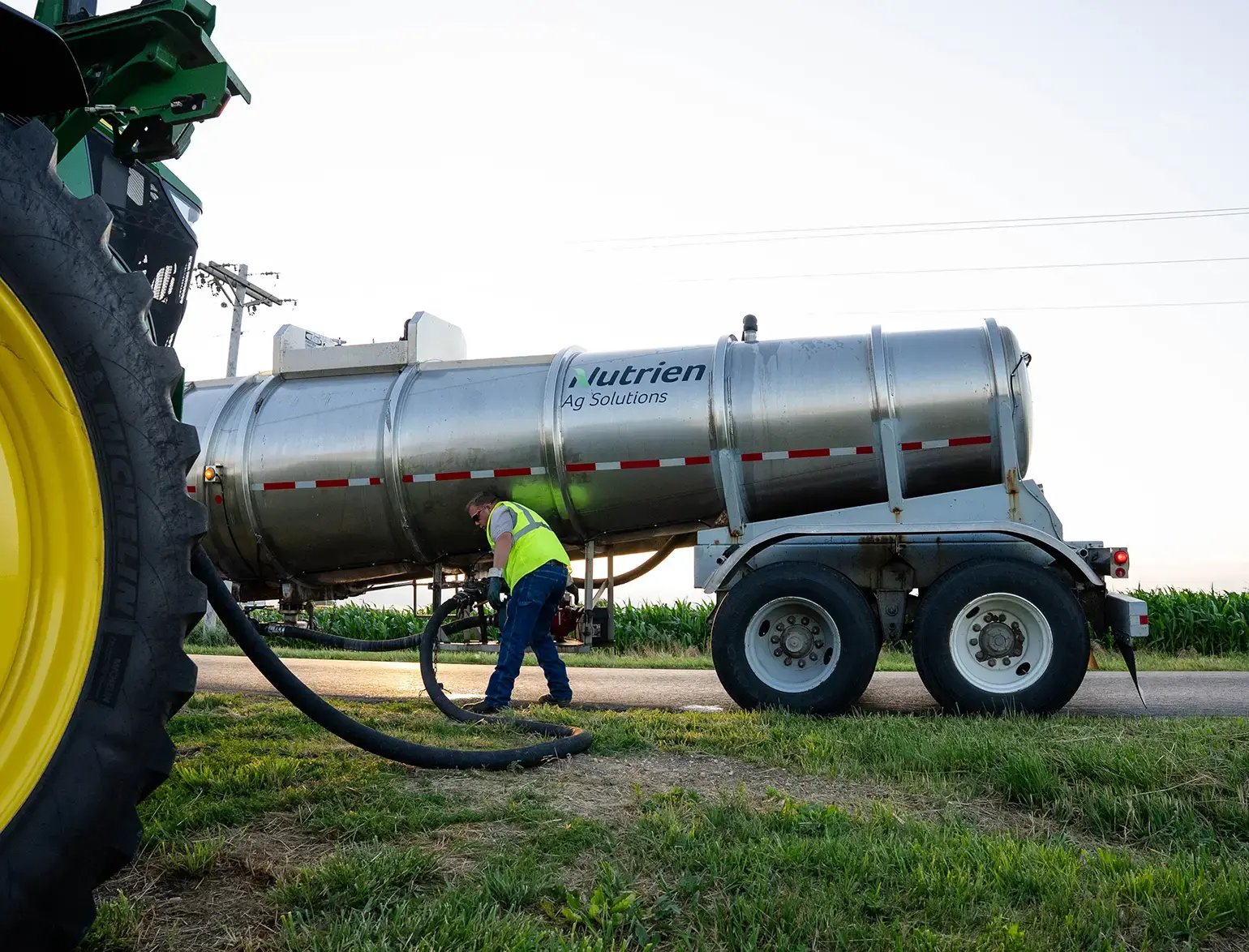 Worker connecting equipment to a Nutrien Ag Solutions tank trailer beside a large farm vehicle.